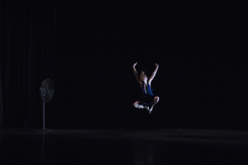 Nicole Wolcott leaps into the air with her legs tucked under the body. A fan is on the stage